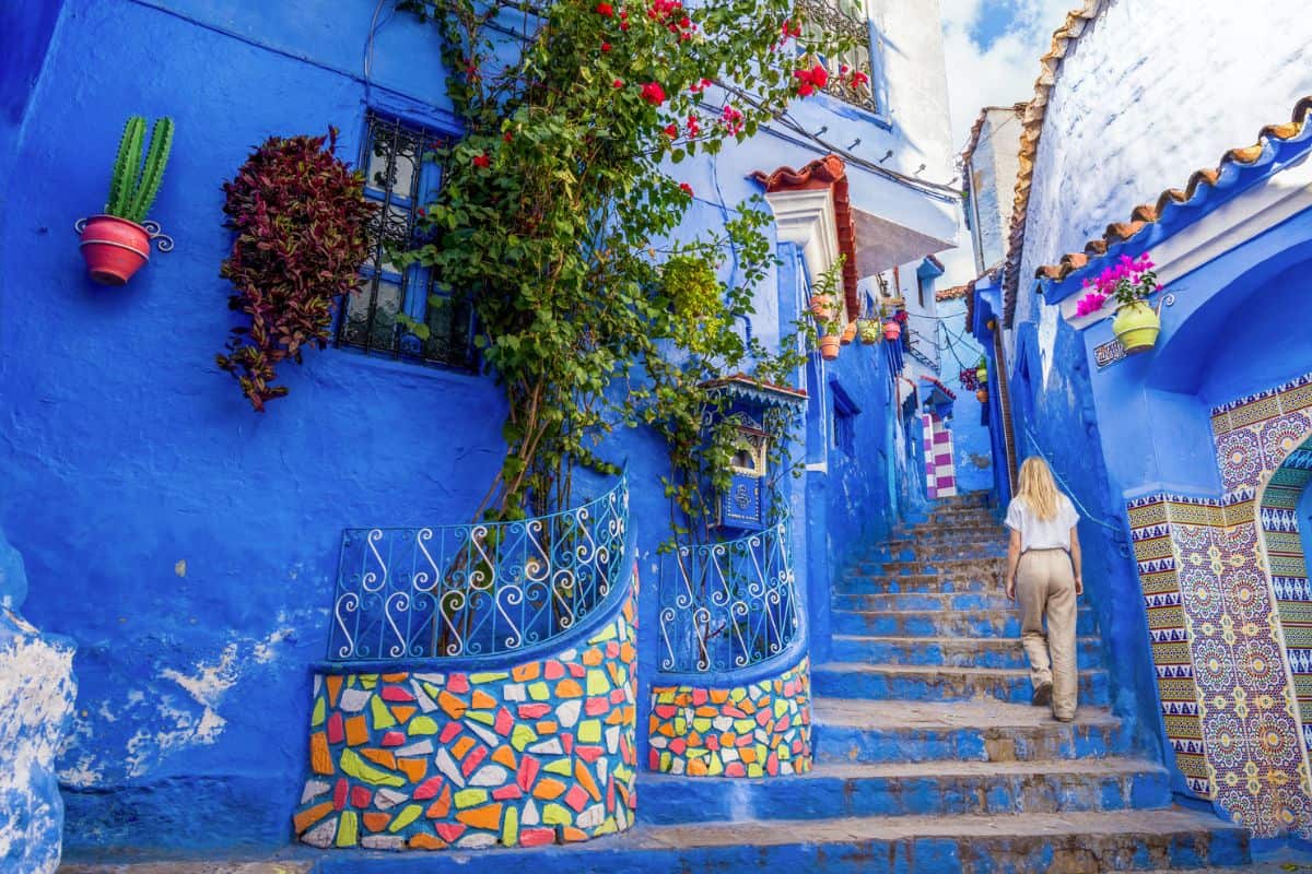 Blick auf eine malerische, blaue Gasse mit bunten Blumen und einer Treppe, die zu einem oberen Bereich führt, umgeben von traditioneller Architektur.