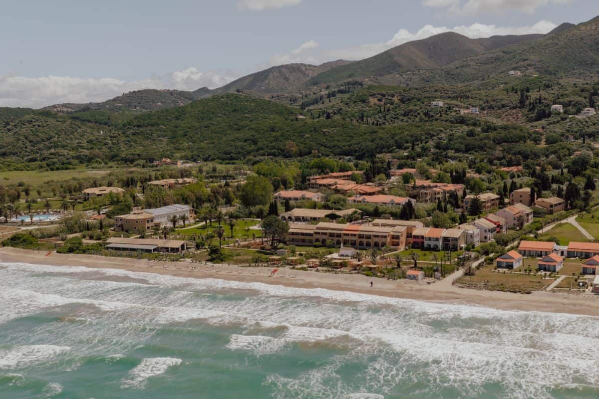 Luftaufnahme eines Küstenresorts mit mehreren Gebäuden, umgeben von grünen Hügeln und einem Sandstrand mit Wellen.