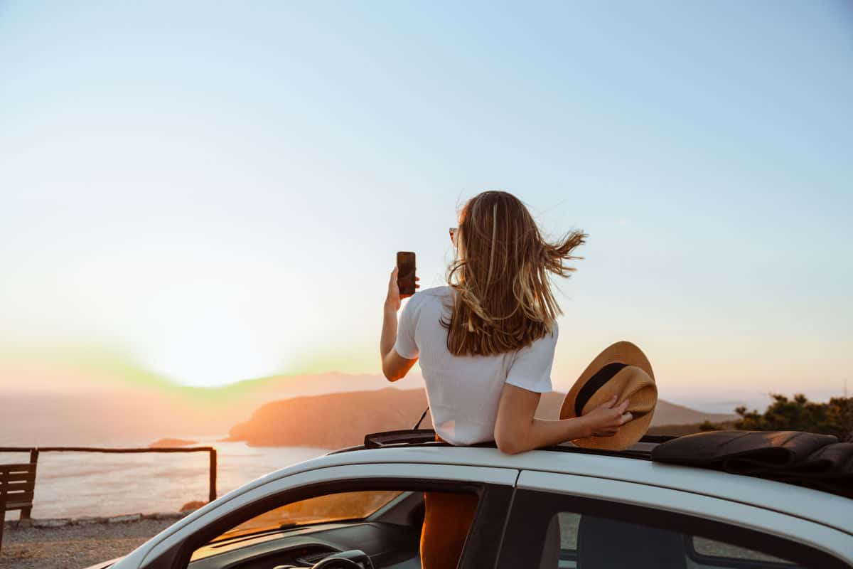 Eine Person sitzt auf dem Dach eines Autos und fotografiert den Sonnenuntergang über dem Meer mit einem Smartphone.