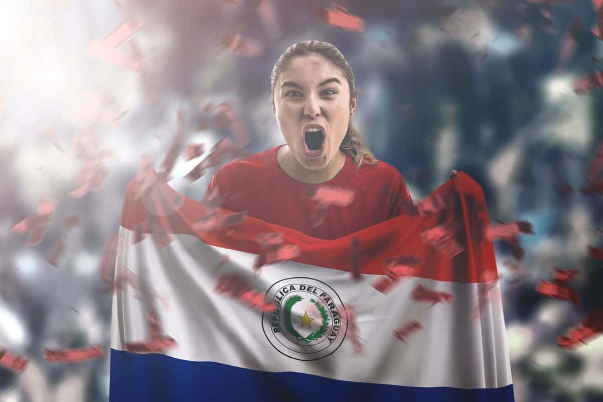 Eine junge Frau in einem roten Shirt hält die Flagge von Paraguay und schreit enthusiastisch, umgeben von Konfetti und einer jubelnden Menge.