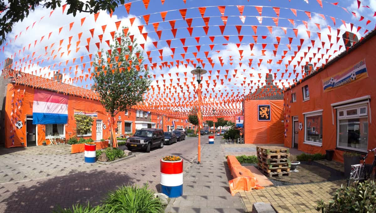 Blick auf eine festlich dekorierte Straße mit orangefarbenen Fahnen und Banner, umgeben von Häusern und Bäumen unter blauem Himmel.
