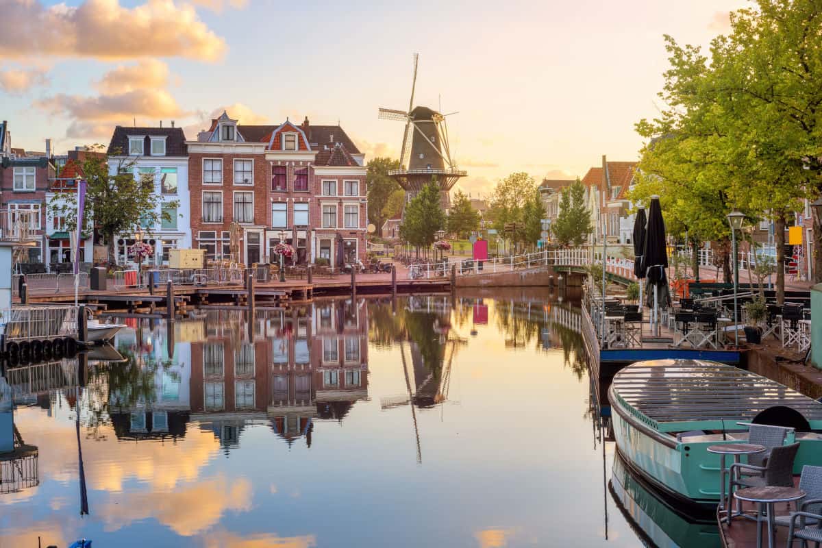 Ein malerischer Kanal mit historischen Gebäuden, einer Windmühle und reflektierendem Wasser, umgeben von Bäumen und Sitzbereichen in der niederländischen Stadt Leiden.