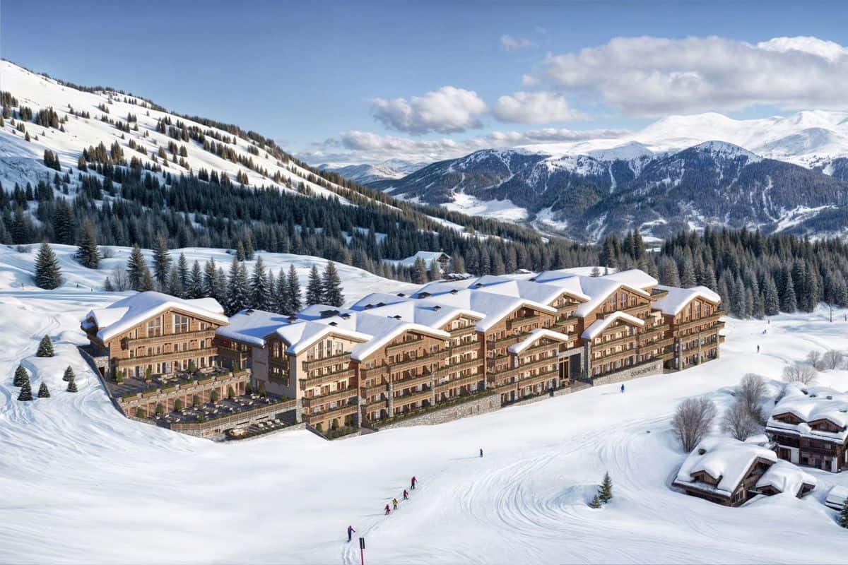 Luftaufnahme eines modernen Hotels in den Alpen, umgeben von schneebedeckten Bergen und Nadelbäumen, mit Skifahrern im Vordergrund.