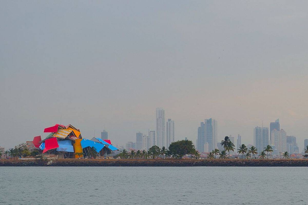 Bunte, moderne Architektur am Wasser mit einer Skyline im Hintergrund, umgeben von Palmen und einem bewölkten Himmel.