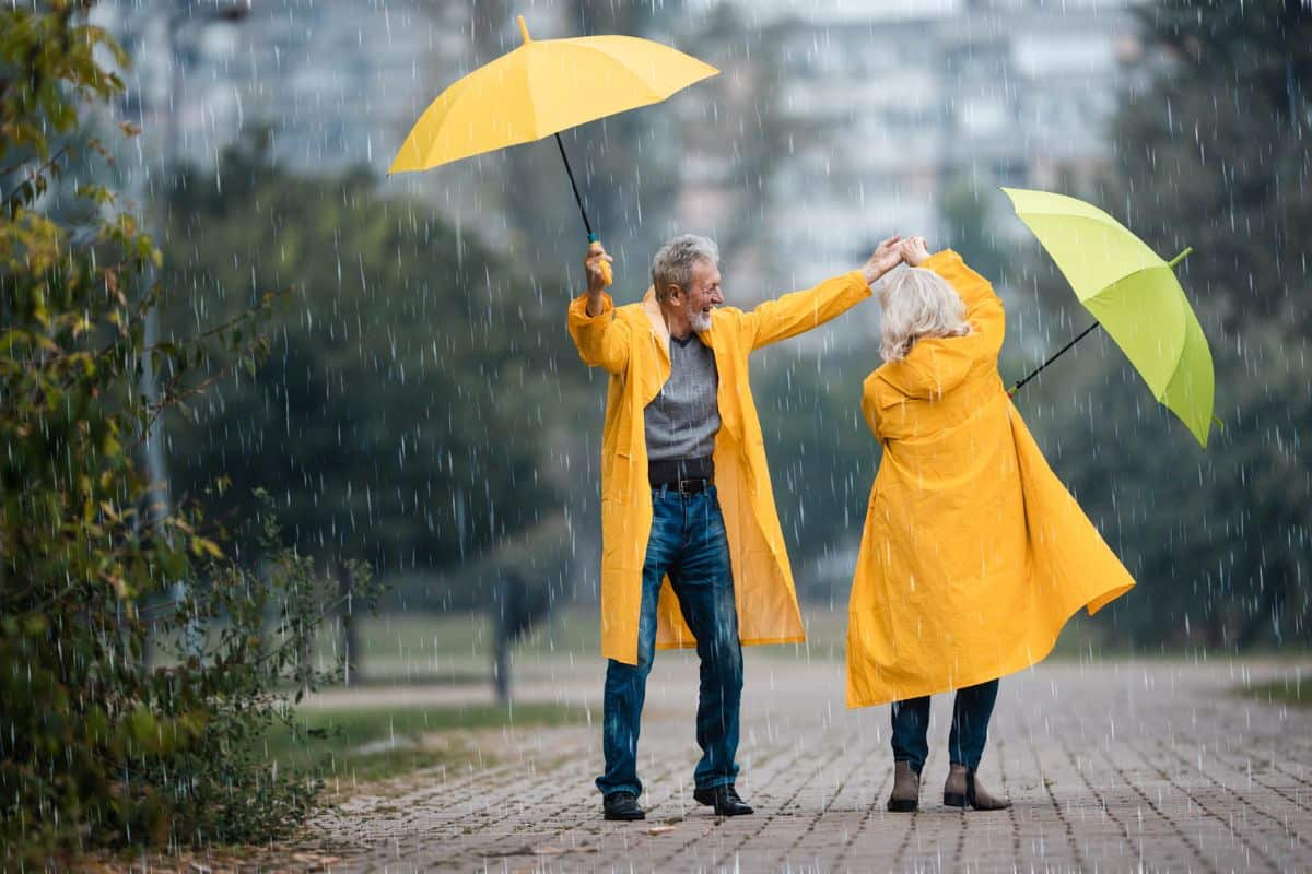 Ein älteres Paar tanzt im Regen, beide tragen gelbe Regenmäntel und halten Regenschirme, während sie auf einem gepflasterten Weg stehen.