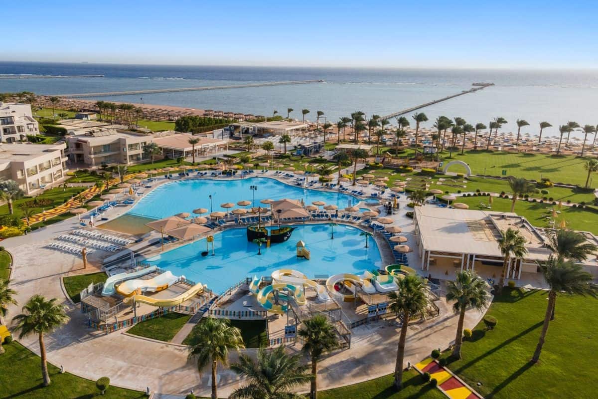 Luftaufnahme eines Resorts mit einem großen Pool, Liegen und Palmen, umgeben von einem Strand und einem Steg, der ins Wasser führt.