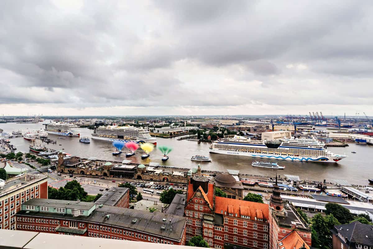 Blick auf den Hafen mit mehreren Kreuzfahrtschiffen, bunten Wasserfontänen und einer städtischen Umgebung unter bewölktem Himmel.
