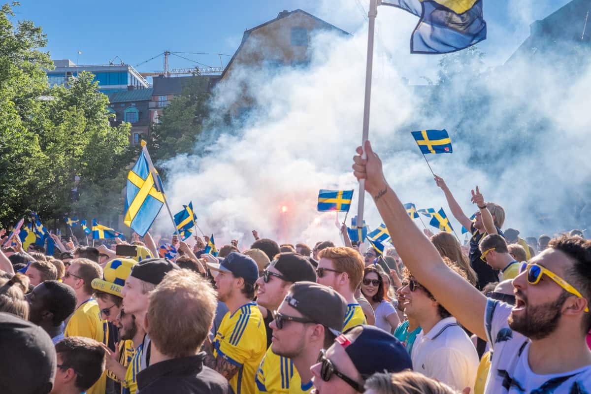 Eine große Menschenmenge von schwedischen Fußballfans feiert im Freien, umgeben von Rauch und schwenkt schwedische Flaggen. Sonnenschein und fröhliche Gesichter prägen die Szene.