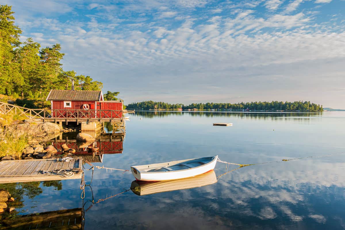 Ruhige Wasserlandschaft mit einem roten Holzhaus, einem kleinen Boot und einer spiegelnden Oberfläche, umgeben von Bäumen und sanften Hügeln.