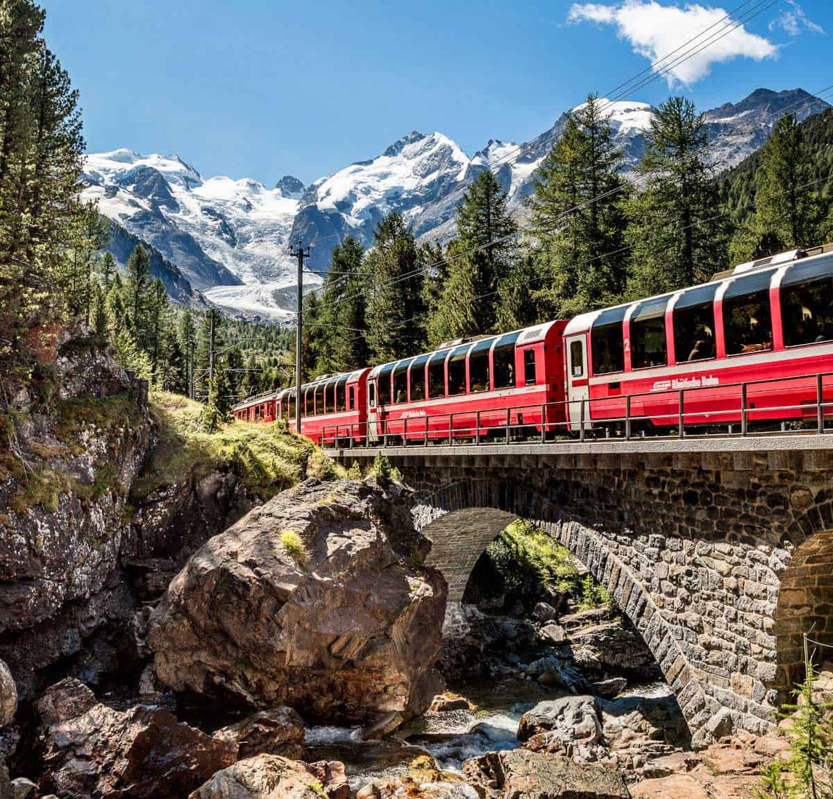 Ein roter Zug fährt über eine Steinbrücke, umgeben von grünen Bäumen und majestätischen Bergen mit schneebedeckten Gipfeln im Hintergrund.