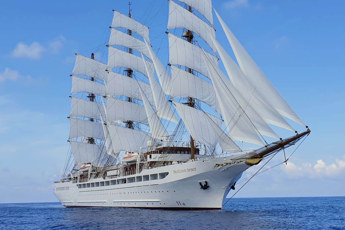 Ein elegantes Segelschiff mit mehreren weißen Segeln auf ruhiger See, umgeben von blauem Himmel und sanften Wolken.