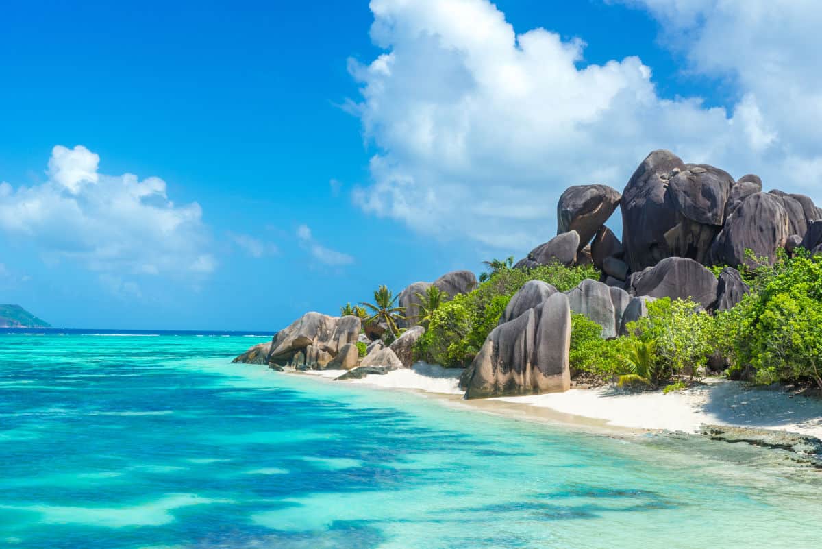 Tropische Küstenlandschaft mit großen Granitfelsen, klarem Wasser und feinem Sandstrand unter einem strahlend blauen Himmel.