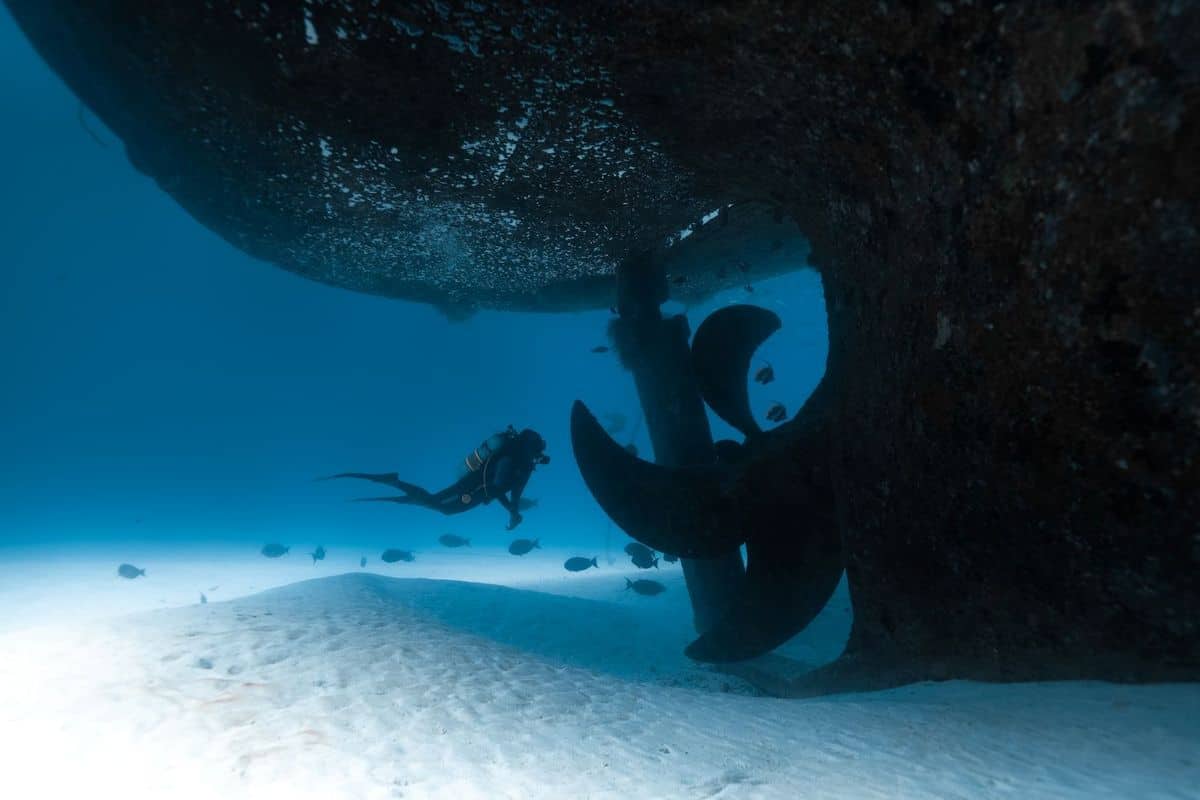 Ein Taucher erkundet den Bereich unter einem Schiffspropeller in klarem Wasser, umgeben von Sand und kleinen Fischen.