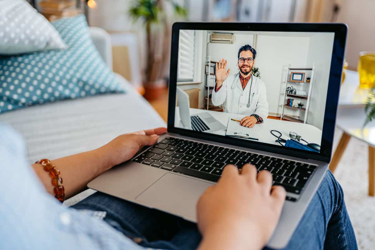 Eine Person sitzt auf einem Sofa und nutzt einen Laptop für eine Videokonferenz mit einem Arzt, der freundlich winkt.