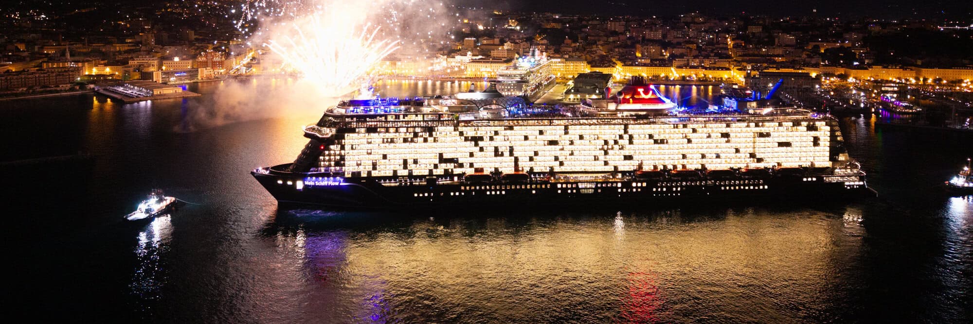 Ein Kreuzfahrtschiff im Hafen, beleuchtet von bunten Lichtern, während Feuerwerk den Nachthimmel erhellt. Reflektionen im Wasser sind sichtbar.