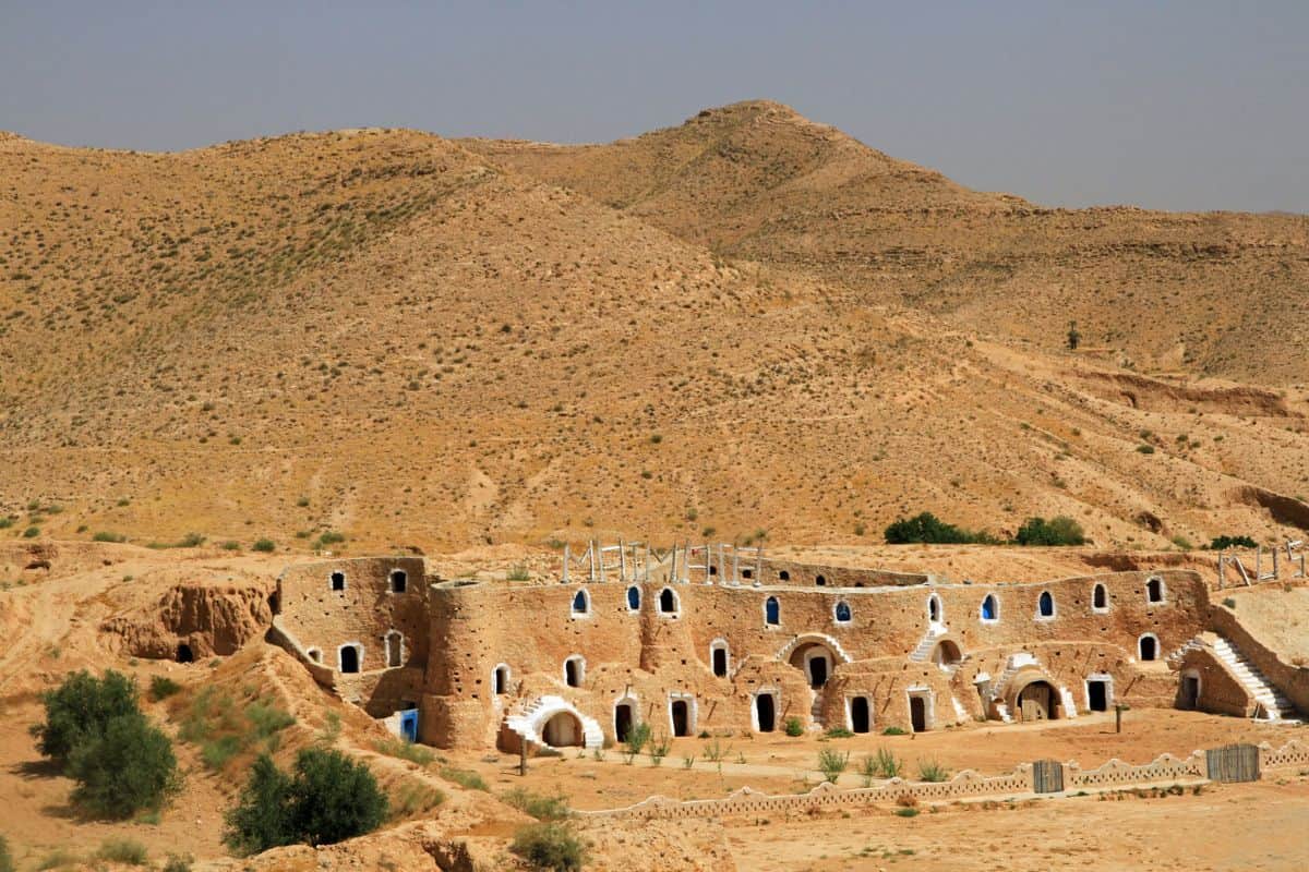 Eine historische, erdige Struktur mit blauen Fenstern vor einer kargen, bergigen Landschaft in der Wüste. Die Umgebung ist trocken und sandig.