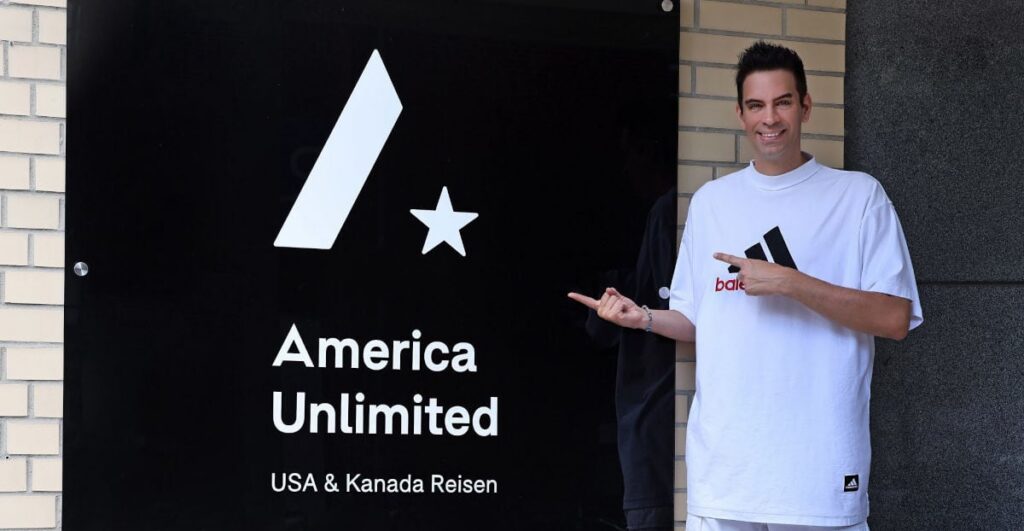 Ein Mann in einem weißen T-Shirt steht vor einem schwarzen Schild mit dem Schriftzug 'America Unlimited' und zeigt mit dem Finger darauf.