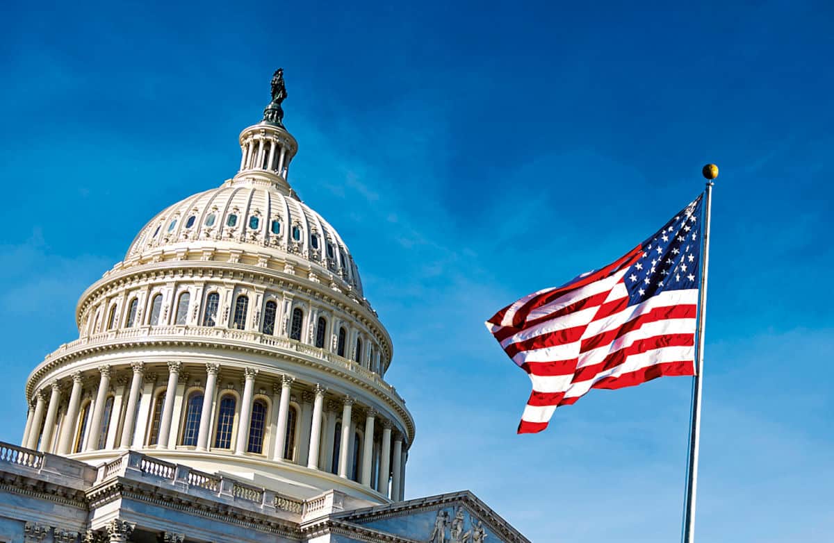 Die Kuppel des Kapitols in Washington D.C. mit einer wehenden amerikanischen Flagge im Vordergrund vor einem klaren blauen Himmel.