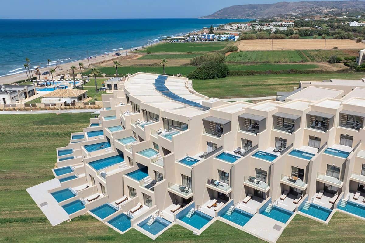Luftaufnahme eines modernen Resorts mit mehreren Zimmern, die über private Pools verfügen, umgeben von grünen Wiesen und dem Meer im Hintergrund.