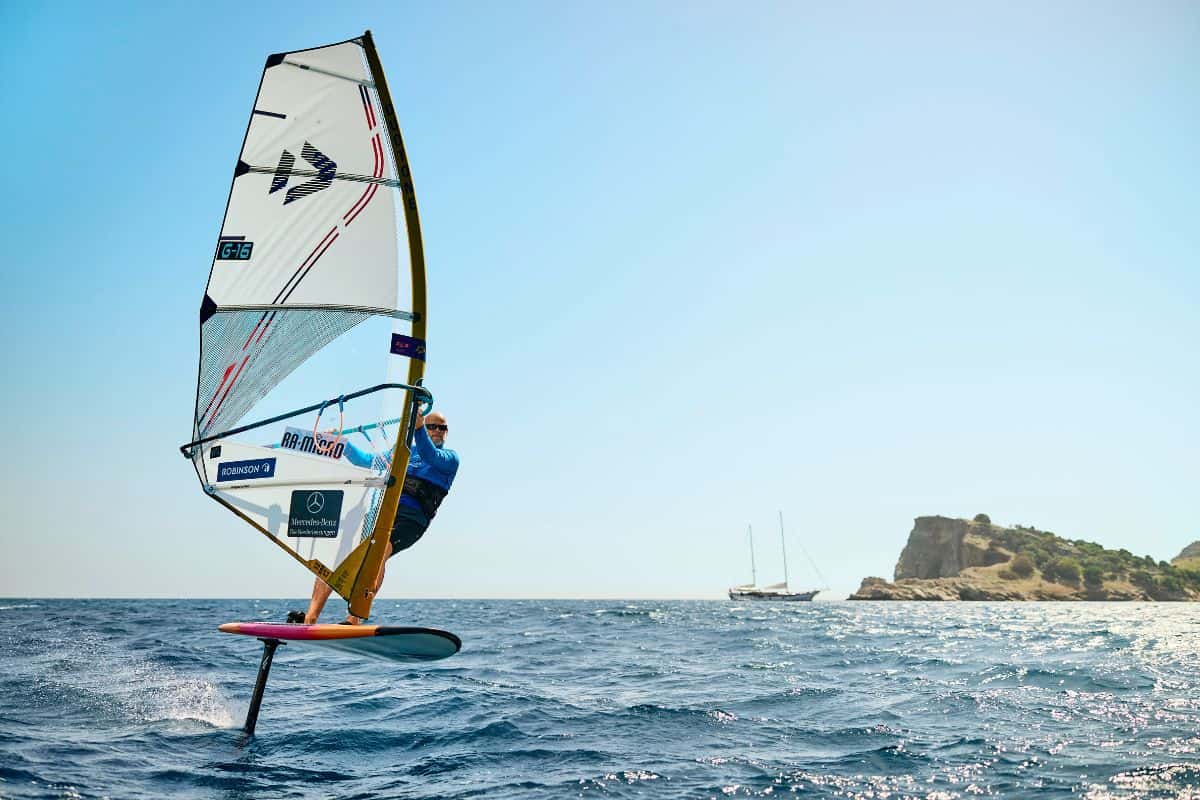Ein Windsurfer gleitet über das Wasser, während im Hintergrund ein Segelboot und eine Küstenlandschaft sichtbar sind. Klare Himmel und ruhige See.