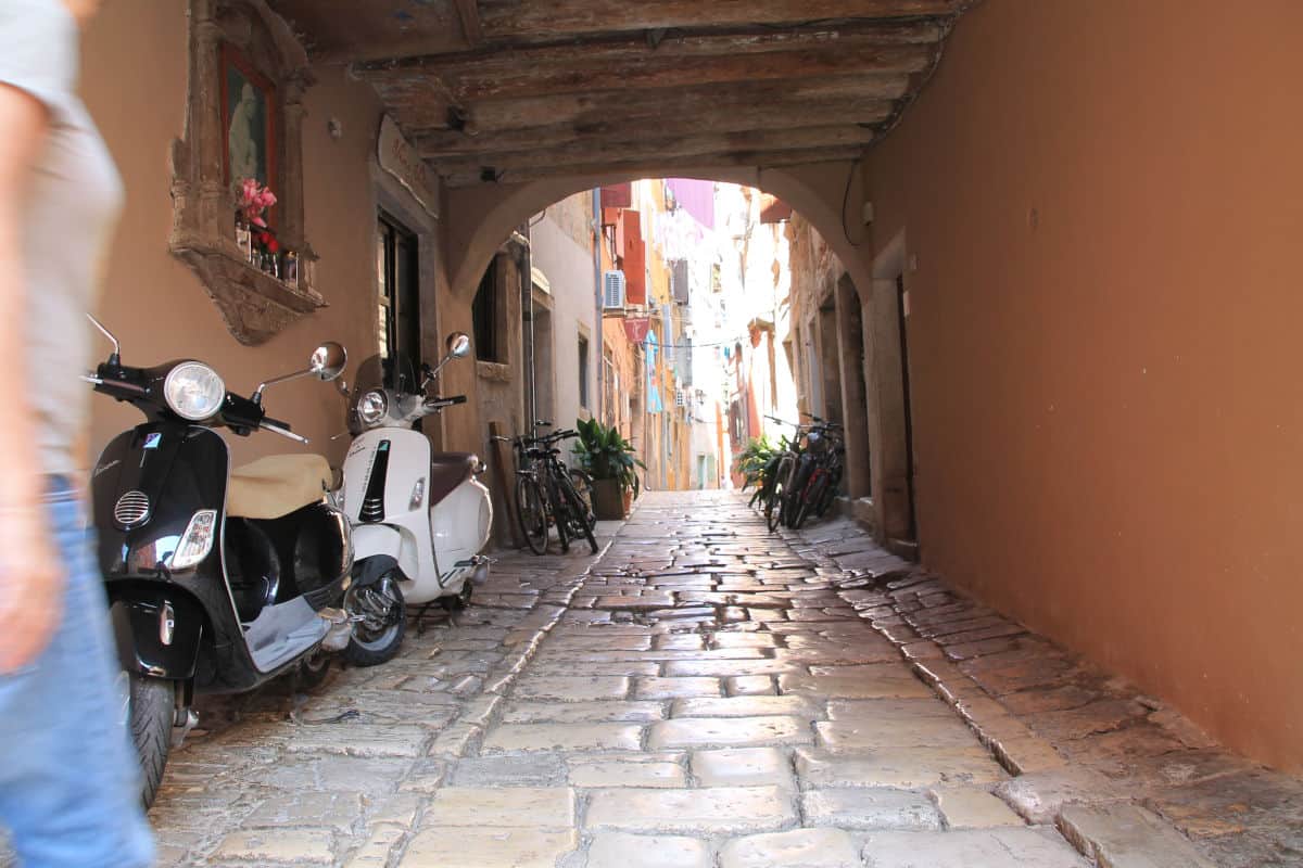 Engpass in einer historischen Gasse mit Mofas und Fahrrädern, umgeben von alten Gebäuden und einem gepflasterten Boden.