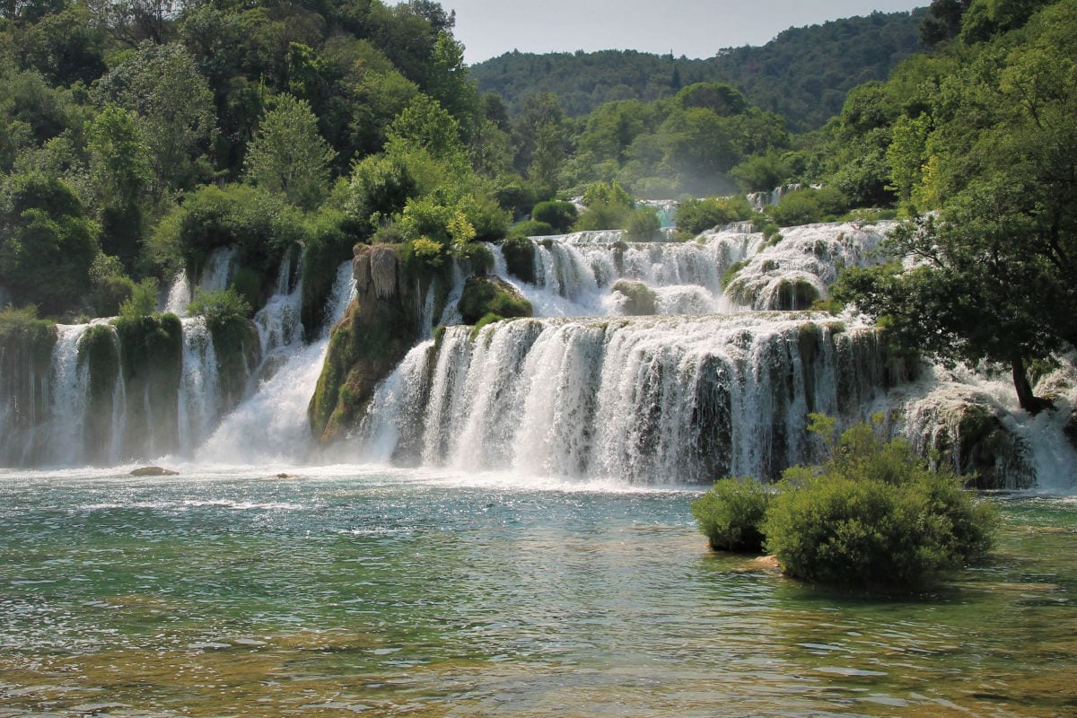 Ein malerischer Wasserfall fließt über mehrere Stufen in einen klaren, ruhigen See, umgeben von üppigem Grün und Bäumen.