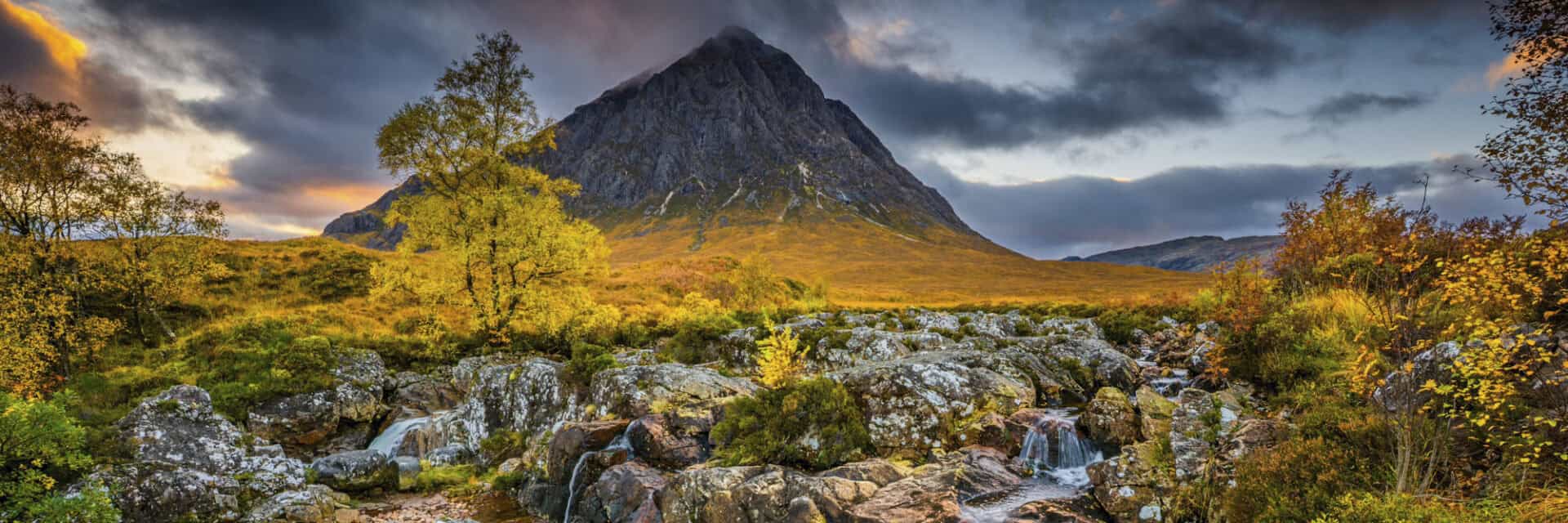 wm-serie-schottland Eine malerische Landschaft mit einem hohen Berg im Hintergrund, umgeben von bunten Bäumen und einem kleinen Wasserfall, der über Steine fließt.