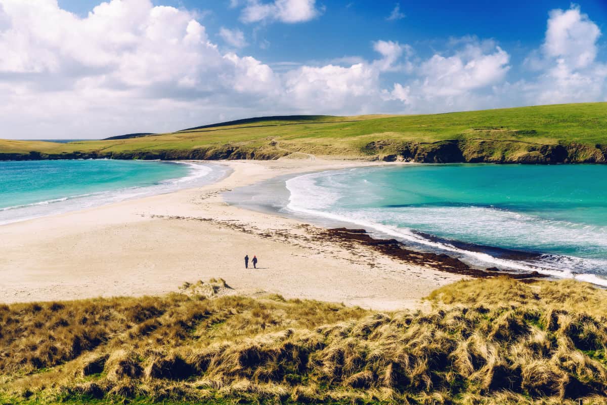 Zwei Personen spazieren an einem weitläufigen Strand mit feinem Sand und klarem, türkisfarbenem Wasser unter einem blauen Himmel mit Wolken.