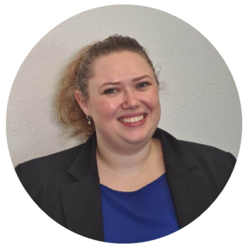 Eine lächelnde Frau mit lockigem Haar, die einen schwarzen Blazer und ein blaues Oberteil trägt, vor einer hellen Wand.