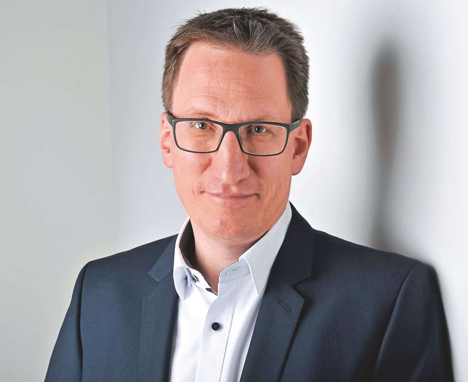 Porträt eines Mannes mit Brille, der in einem Anzug gekleidet ist. Er steht vor einer hellen Wand und lächelt leicht.