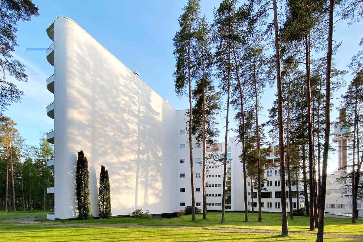 Ein weißes, geschwungenes Hochhaus mit Balkonen steht in einer bewaldeten Parklandschaft mit hohen Kiefern unter einem klaren, hellblauen Himmel bei Tag.