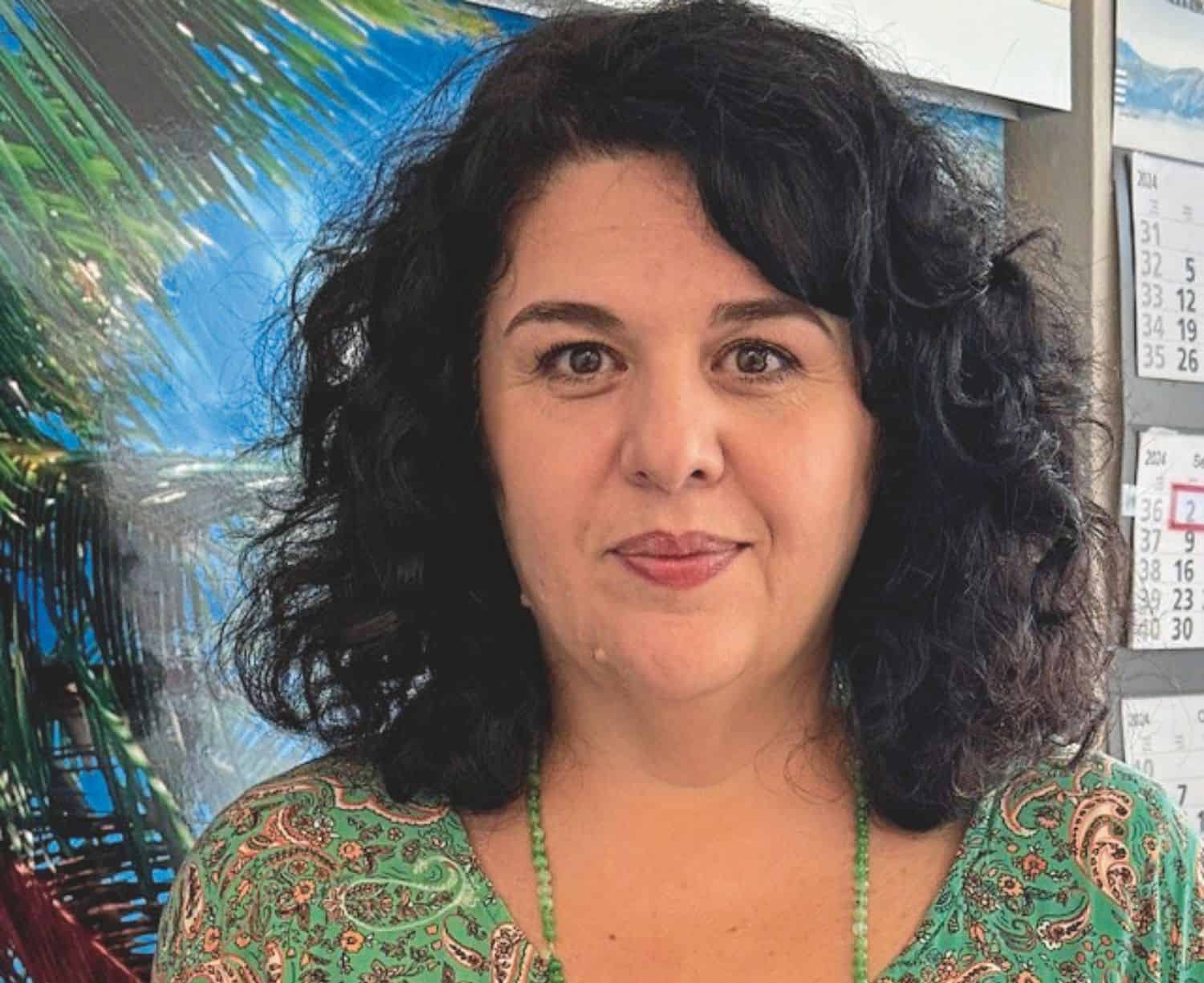 Porträt einer Frau mit lockigem, schwarzem Haar, die ein grünes, gemustertes Oberteil trägt, vor einem Hintergrund mit Palmen und einem Kalender.