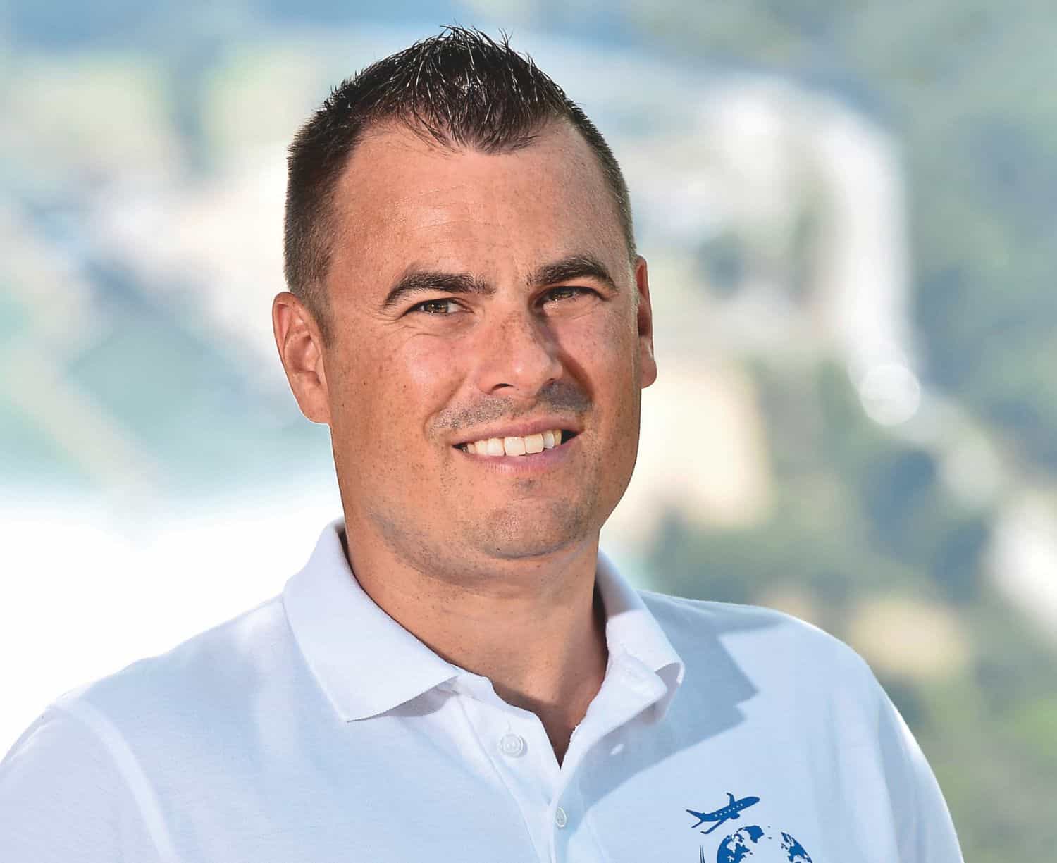 Porträt eines Mannes mit kurzen, dunklen Haaren, der ein weißes Poloshirt trägt, vor einer unscharfen, natürlichen Hintergrundlandschaft.
