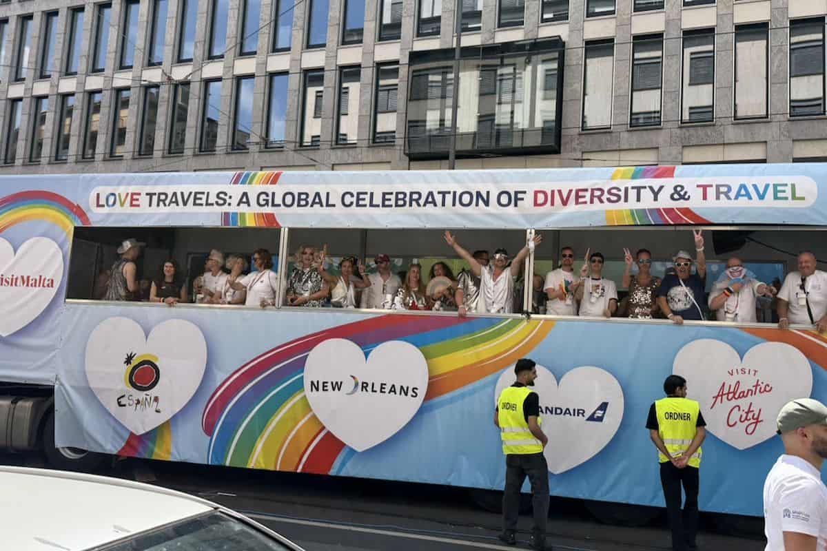 Ein bunter Reisebus mit Regenbogen-Design und verschiedenen Logos fährt an einem modernen Bürogebäude vorbei, während Menschen auf dem Oberdeck winken.