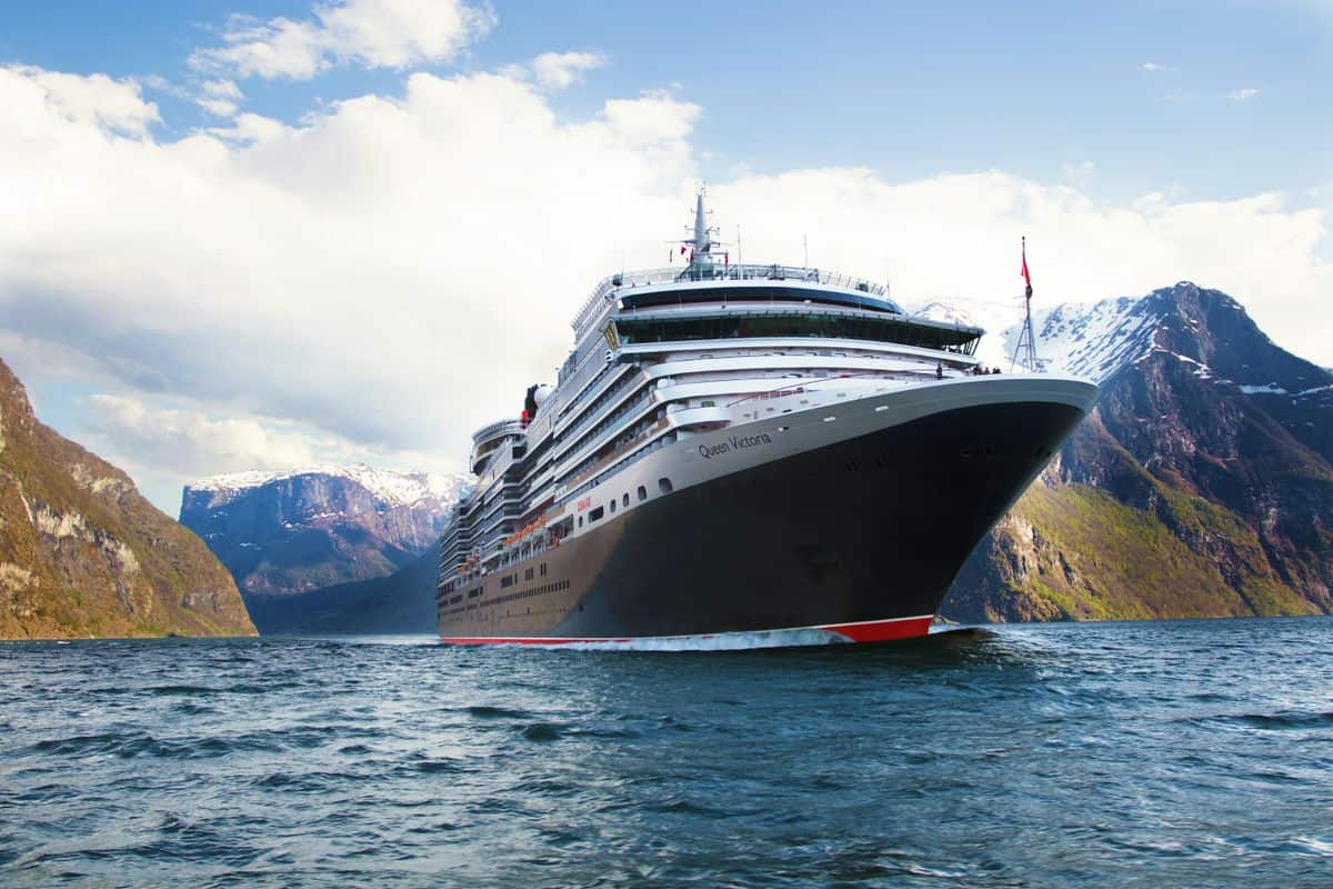 Das Kreuzfahrtschiff Queen Victoria fährt durch einen tiefen Fjord, der von hohen, schneebedeckten Bergen und blauem Himmel mit hellen Wolken umgeben ist.