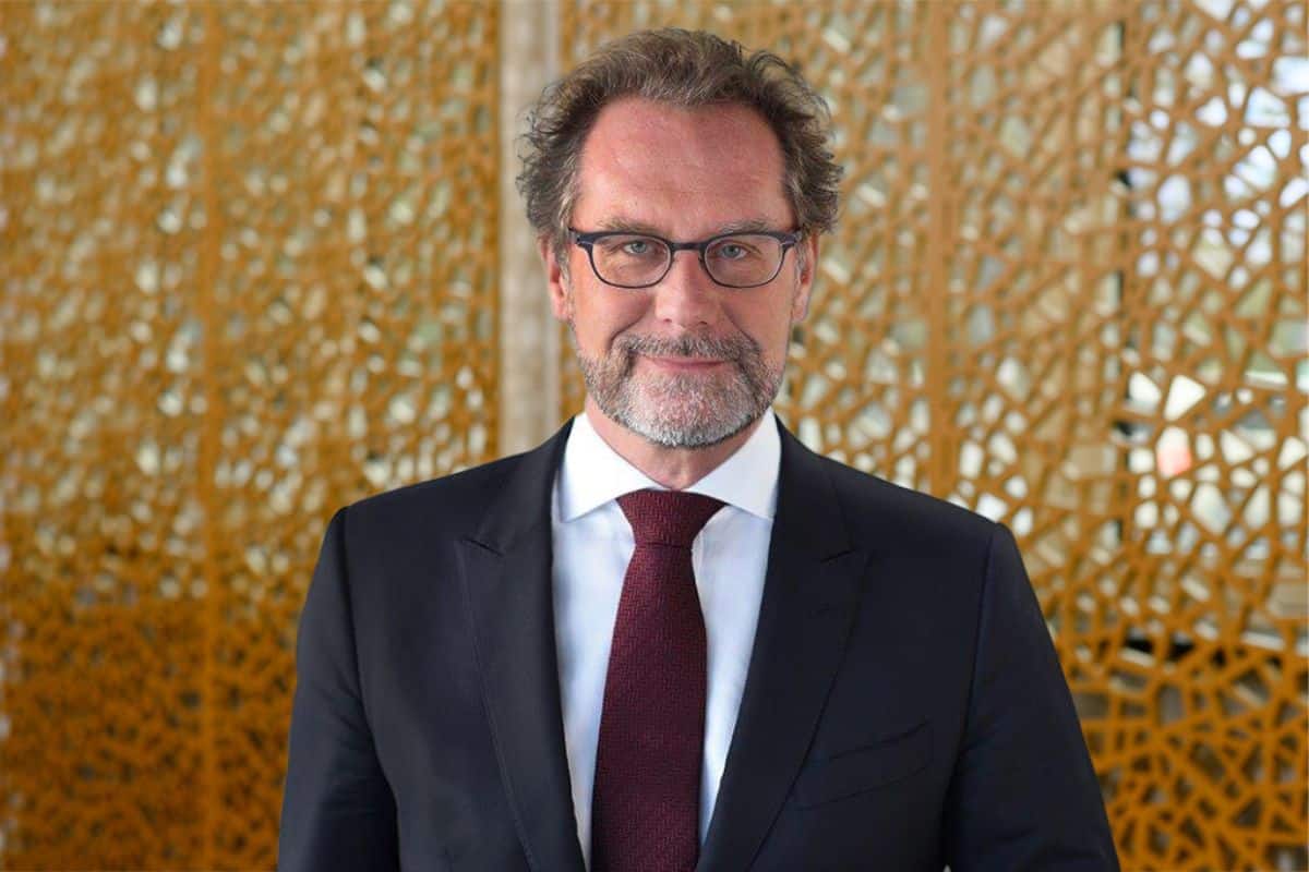 Ein Mann mittleren Alters mit Brille, Vollbart und dunklem Anzug mit bordeauxroter Krawatte vor einem goldfarbenen, ornamentalen Hintergrund im Porträt.