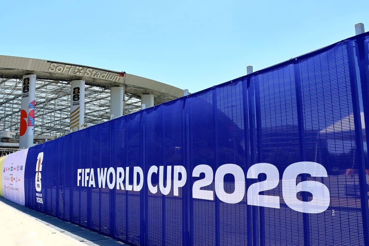 Ein blauer Bauzaun mit weißem FIFA World Cup 2026 Schriftzug steht vor dem SoFi Stadium, an dessen Säulen Banner mit dem Turnierlogo befestigt sind.