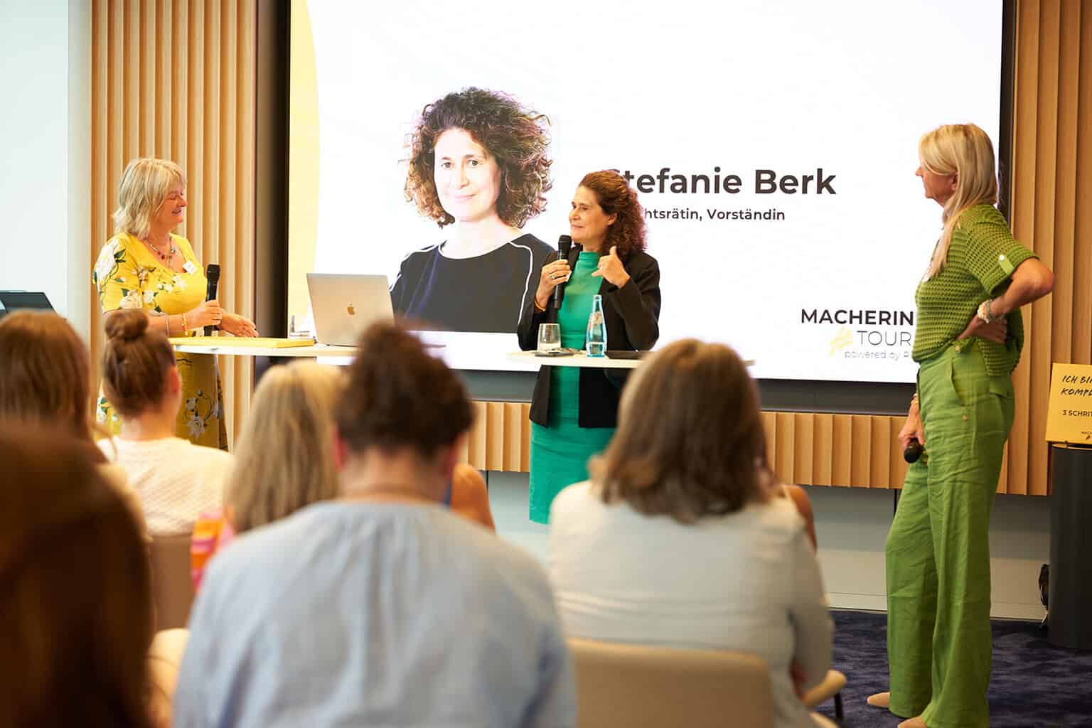 Drei Frauen stehen an einem Tisch während einer Präsentation. Im Hintergrund ist ein Bildschirm mit einem Bild und Text zu sehen.