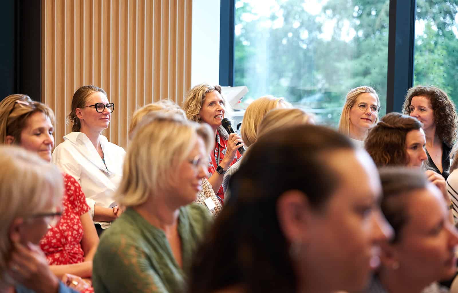 Eine Gruppe von Frauen sitzt in einem modernen Raum, einige hören zu, während eine Frau mit Mikrofon spricht. Helle Farben und große Fenster sind sichtbar.