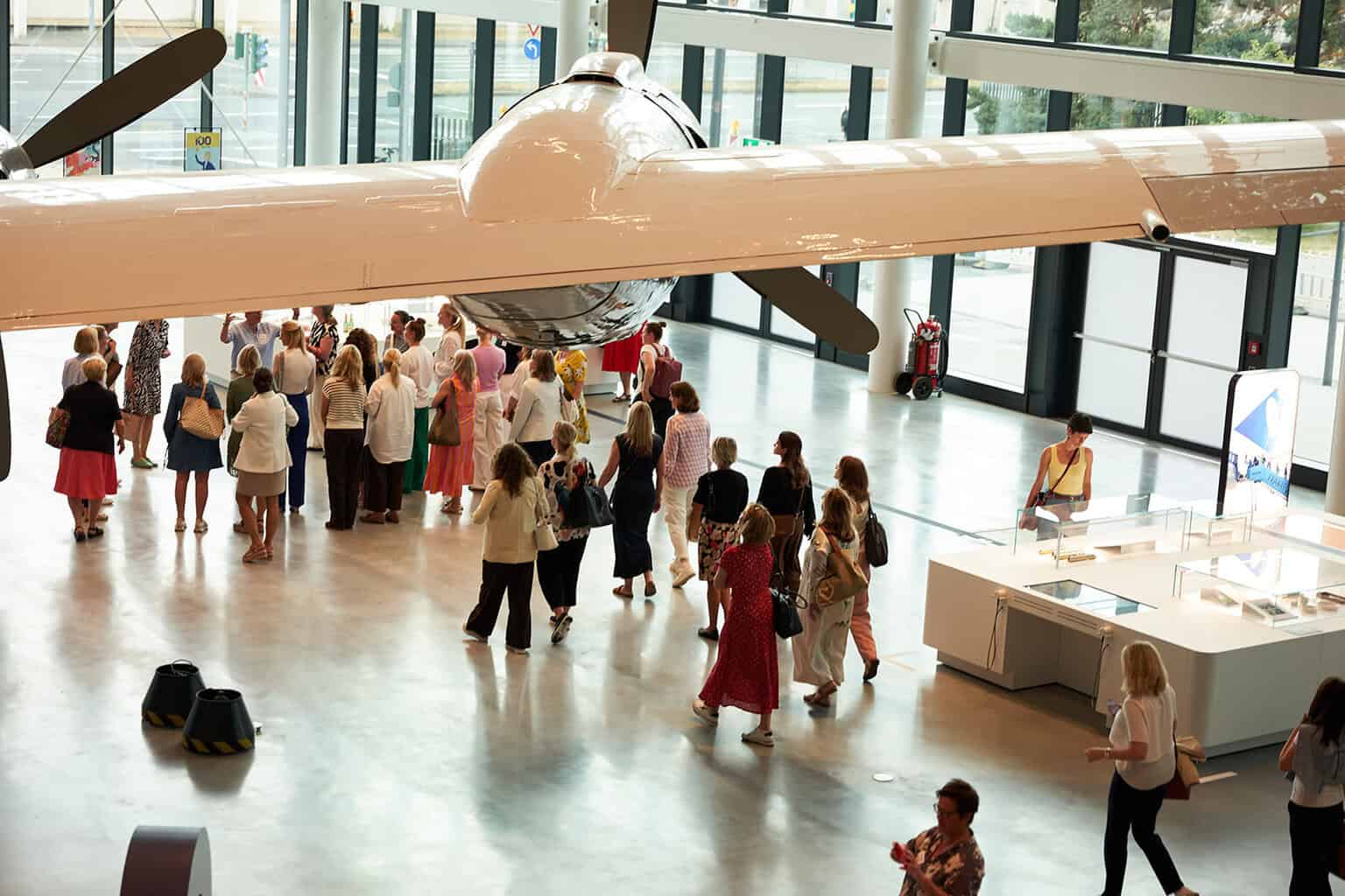 Eine Gruppe von Personen steht in einem modernen Ausstellungsraum unter einem großen Flugzeugmodell. Helle Wände und viel Tageslicht sind sichtbar.