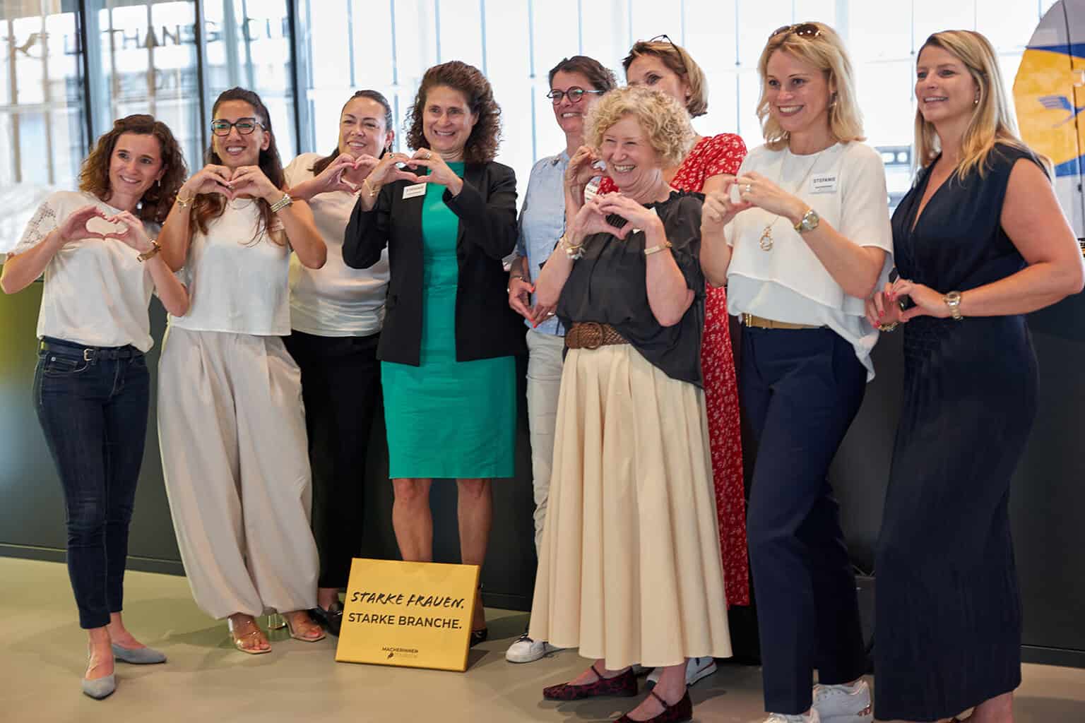 Eine Gruppe von Frauen steht zusammen und formt mit ihren Händen Herzen. Sie tragen verschiedene Outfits und lächeln in die Kamera.