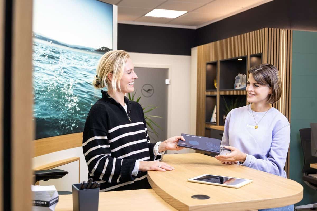 Zwei Frauen stehen an einem hellen Holztresen in einem modernen Büro. Eine Frau in einem gestreiften Pullover überreicht der anderen eine dunkle Dokumentenmappe.