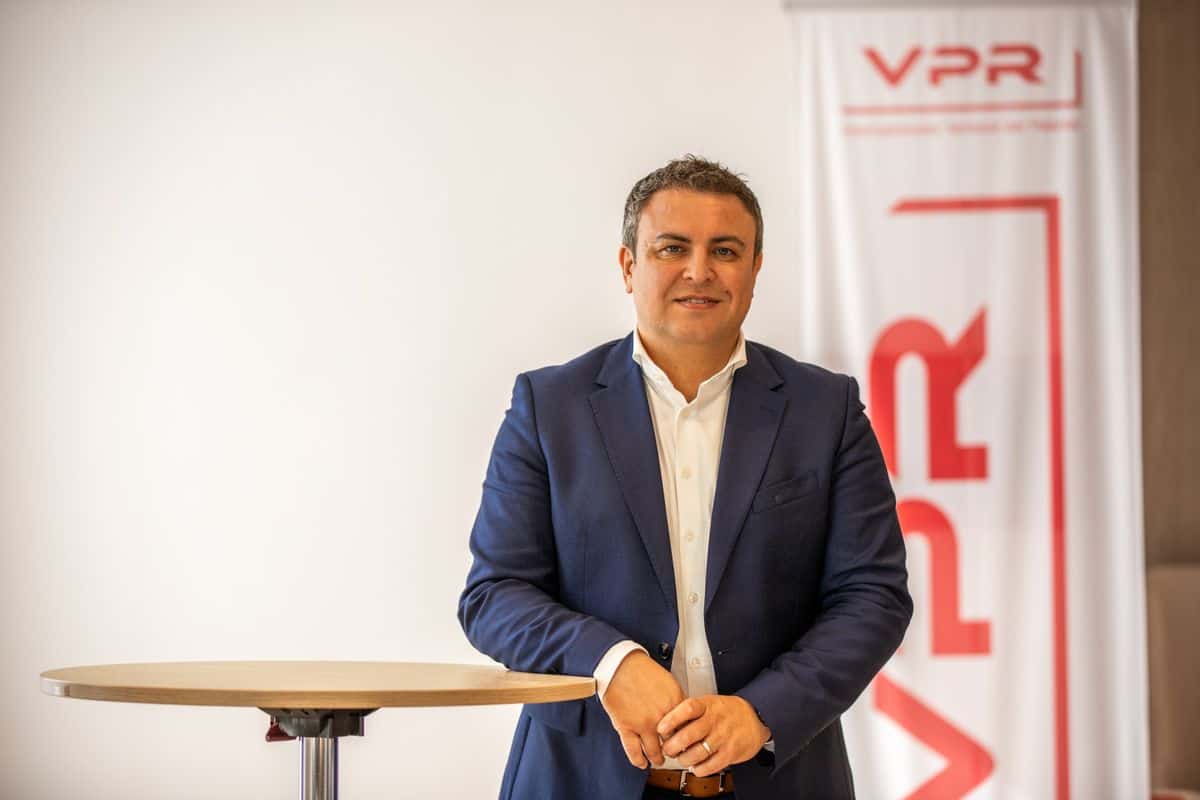 Ein Mann mit kurzem dunklem Haar trägt ein weißes Hemd und ein dunkelblaues Sakko. Er steht vor einem weißen Banner mit rotem VPR-Logo und lächelt leicht.