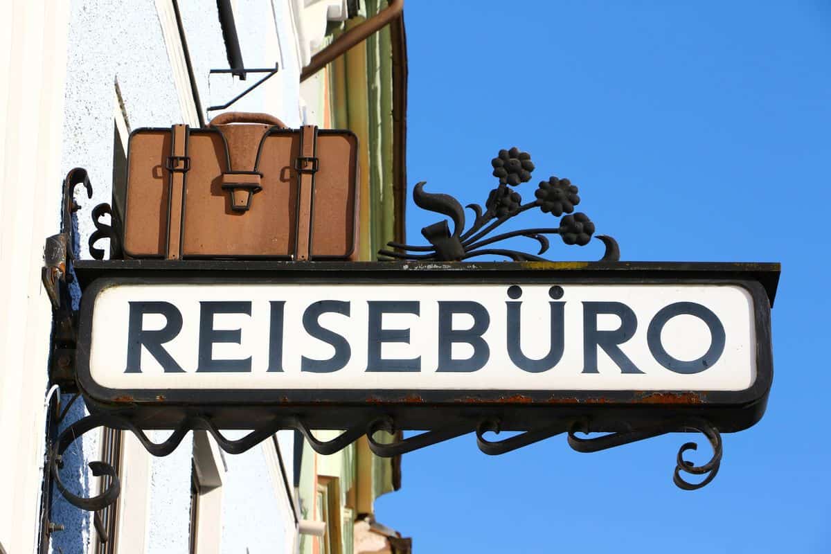 Ein dekoratives Schild mit der Aufschrift 'REISEBÜRO' und einem alten Koffer darüber, vor einem klaren blauen Himmel.