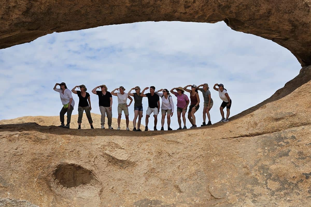 Eine Gruppe von zwölf Personen steht in einer Reihe auf einem Felsen und schaut durch eine natürliche Steinöffnung in den Himmel.