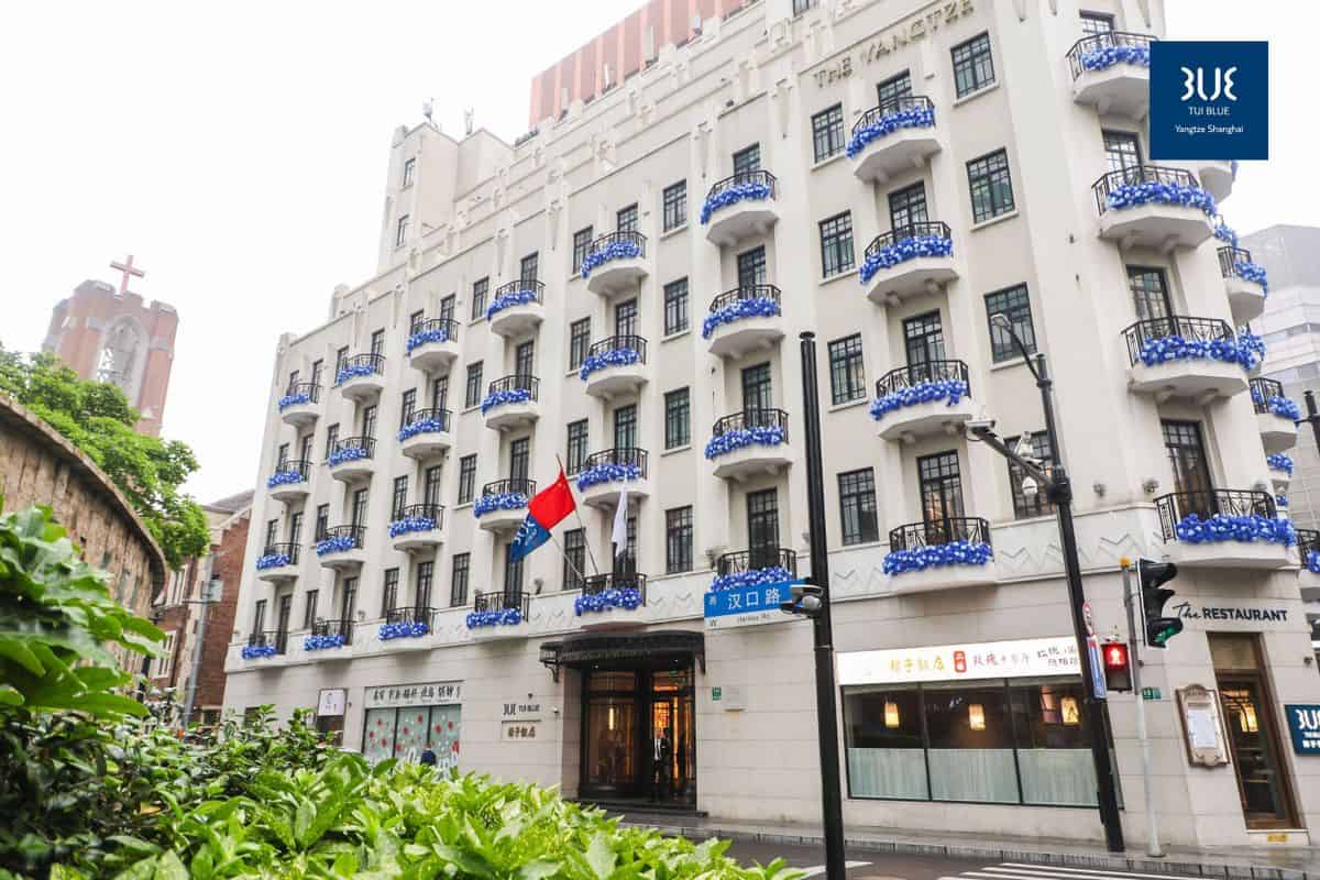 Das Gebäude eines Hotels in Shanghai mit blauen Balkonen und einer chinesischen Flagge. Im Hintergrund ist ein Kirchturm sichtbar.