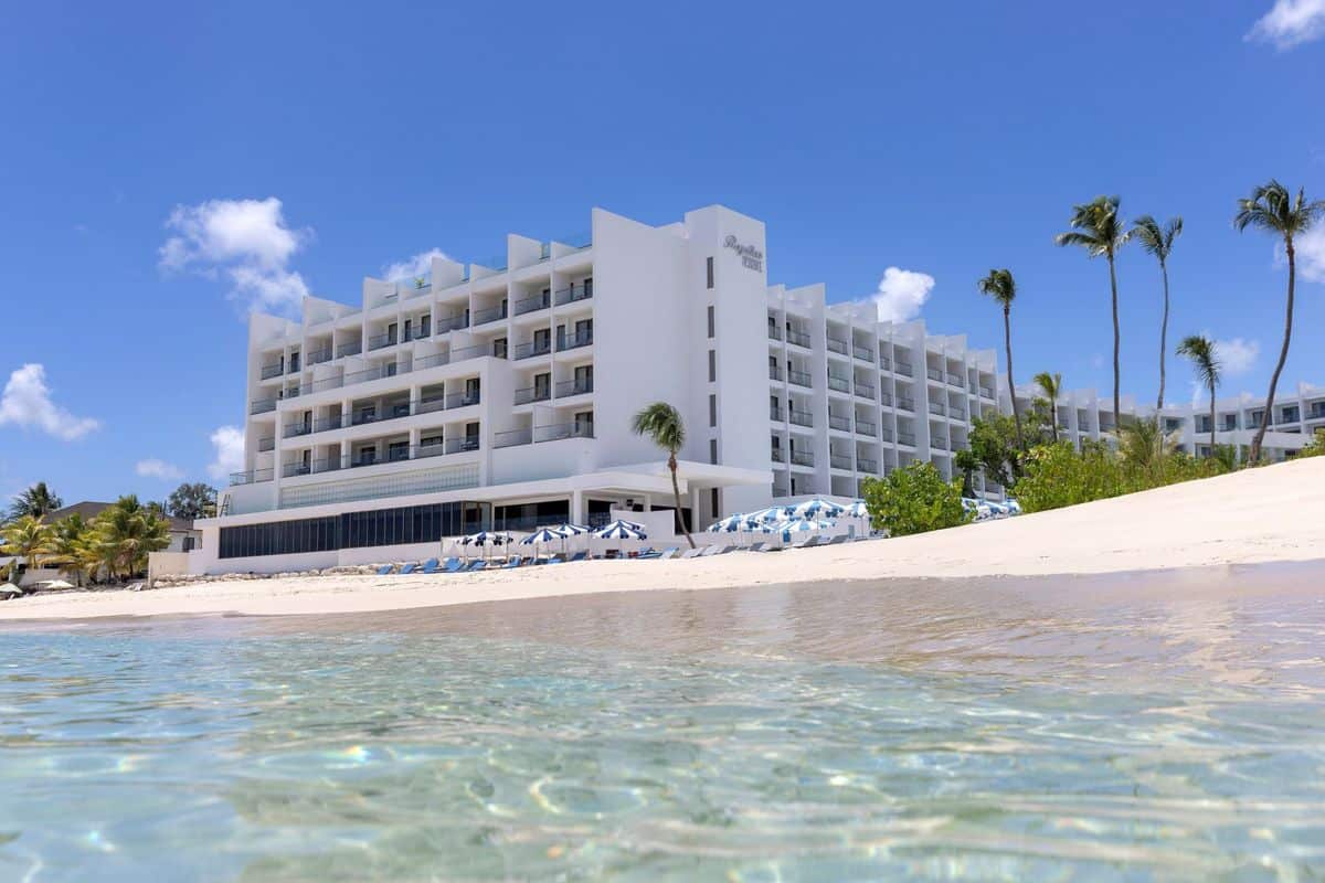 Modernes Hotelgebäude mit mehreren Etagen, umgeben von Palmen und einem weißen Sandstrand, unter einem klaren blauen Himmel.