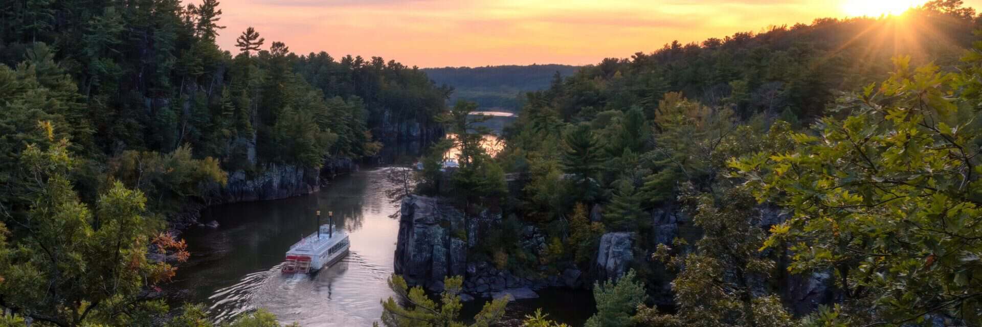 mississippi Ein Raddampfer fährt bei Sonnenuntergang durch eine enge, von bewaldeten Felswänden gesäumte Flusslandschaft, wobei das Licht den Himmel in Goldtöne taucht.