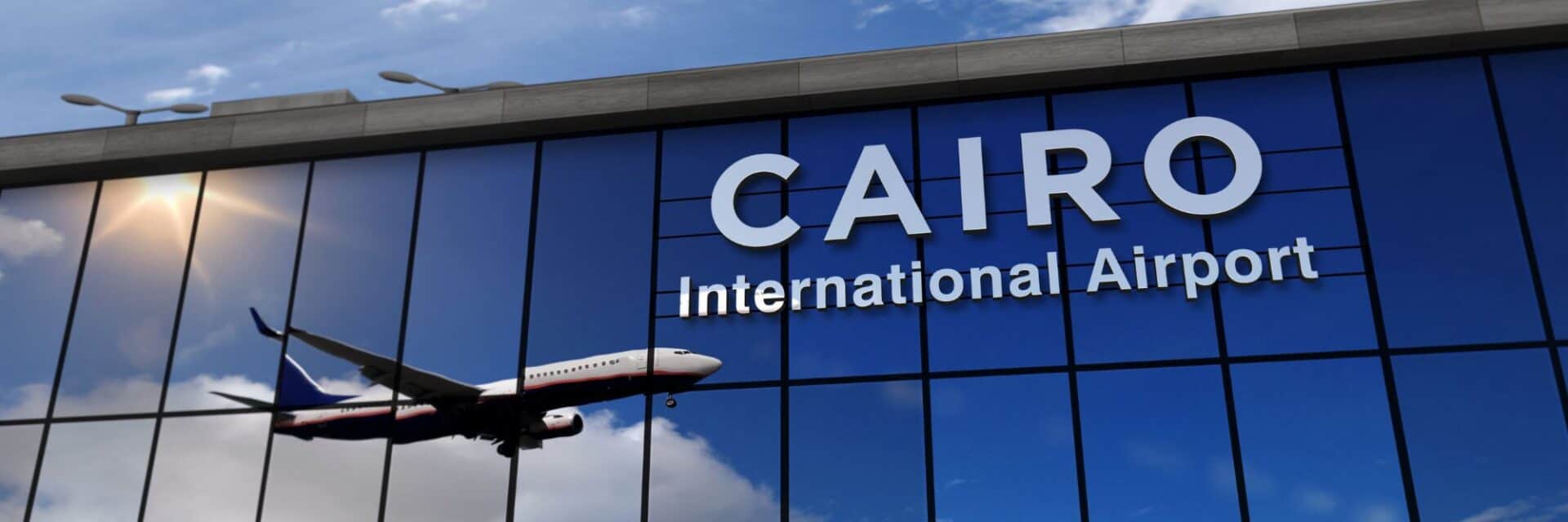 kairo-flughafen-ägypten Die Glasfassade des Cairo International Airport mit dem Schriftzug und der Spiegelung eines startenden Flugzeugs unter einem hellblauen Himmel mit Wolken.