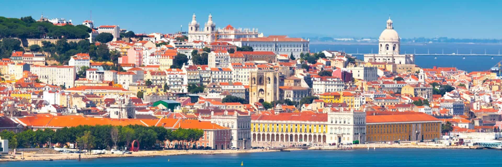 lissabon-tui Weitwinkelaufnahme von Lissabon mit dicht bebauten Häuserfassaden, roten Ziegeldächern, der markanten Kuppel der Panteão Nacional und dem Tejo im Vordergrund.